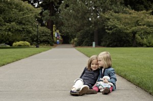 Little blonde girls sitting and hugging on a sidewalk